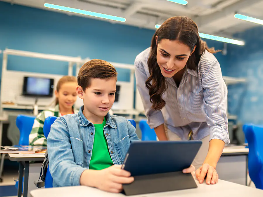 Eine Lehrkraft beugt sich zu einem Jungen und schaut mit ihm auf sein Tablet, im Hintergrund arbeitet ein Mädchen